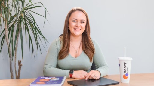 Lauren Roberts, smiling with long brown hair sits at a desk in an office setting. She is wearing a light green top and a red smartwatch, with her hands resting on a closed laptop. On the desk are a smartphone, a purple pen, a spiral notebook with a visible cover, and a branded cup with the logo "SCOTT media." A tall indoor plant is in the background against a light grey wall.