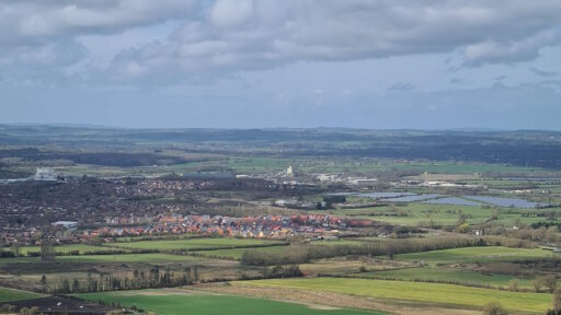 A photograph of the Wiltshire landscape taken from Westbury White Horse showing lots of green space, buildings and a blue sky with some clouds