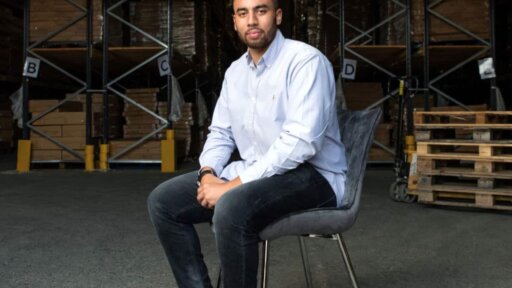Dan Beckles seated on a chair in a warehouse, wearing a light blue shirt, dark jeans, and tan loafers, with stacked pallets and shelving in the background.