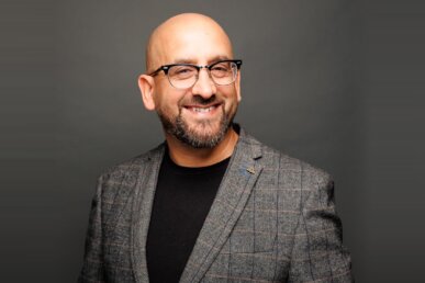 Nathan Sandhu, Founder of Jazzbones Creative, smiling at the camera. He is wearing glasses, a grey check-pattern blazer, and a black shirt, with a neutral grey background.