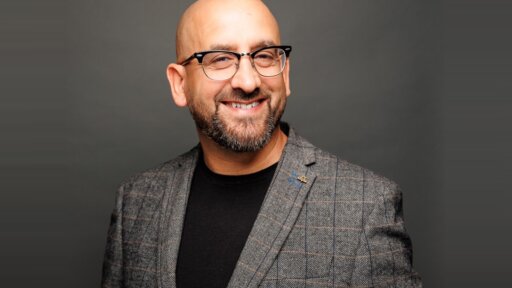 Nathan Sandhu, Founder of Jazzbones Creative, smiling at the camera. He is wearing glasses, a grey check-pattern blazer, and a black shirt, with a neutral grey background.