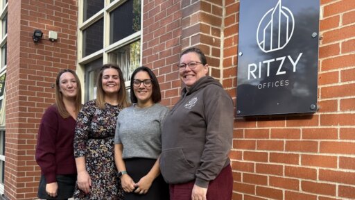 Anna Pawlowska and Rosie Linstead of Ritzy Offices stand alongside Jodie Dallas and Claire Puttick of Sylvian Care in front of a brick building with a sign that reads "RITZY OFFICES." All four individuals are smiling and standing close together, suggesting a friendly and professional collaboration. The building features large windows and a mounted security camera, with a clear blue sky and natural light enhancing the scene.