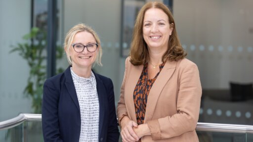 Mary Waters (left) and Jess McHugh (right) stand together in a modern office setting, both dressed in business attire. Mary wears a dark blazer and glasses, while Jess wears a light brown blazer over a patterned dress. They are smiling in front of a glass partition with circular frosted designs, marking their arrival as new Partners in Thrings’ commercial property team.