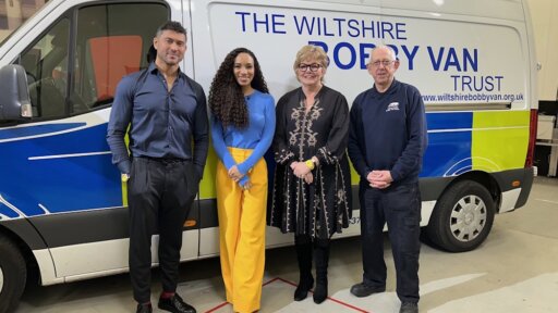 Four people stand smiling in front of a Wiltshire Bobby Van Trust vehicle. From left to right are Ray Wilding in a blue shirt, Michelle Ackerley in a bright blue top and yellow trousers, Jennie Shaw in a black embroidered dress, and Mick Leighfield in uniform.