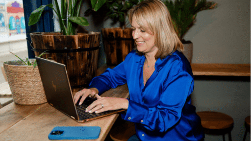 Emma Boatman, Director of Boatman Admin Services, sits at a wooden table in a relaxed indoor setting, smiling while working on a laptop. She wears a vibrant blue satin blouse, with a blue smartphone placed in front of her. The space is bright and welcoming, with large potted plants adding greenery around the table—suggesting a calm, productive atmosphere.