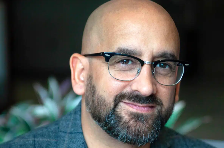 Nathan Sandhu with glasses and a beard wearing a grey blazer. The background is softly blurred with green plants visible.