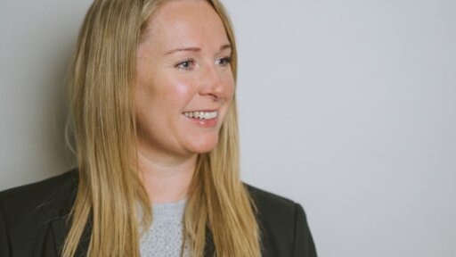Elaine Meyrick, solicitor at Thrings, smiling slightly while looking to the side. She wears a black blazer over a light grey textured top, with long blonde hair against a plain light background.