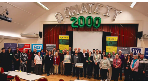 A large group of people standing together in a community hall beneath silver and green balloon letters spelling “DONATE IT 2000.” Behind them are banners for Donate IT, Blackmore UK, and Fixy, along with a projector screen. The setting includes red curtains, tables with chairs in the foreground, and promotional materials on display, indicating a celebratory event marking Donate IT’s 2,000th refurbished device milestone.
