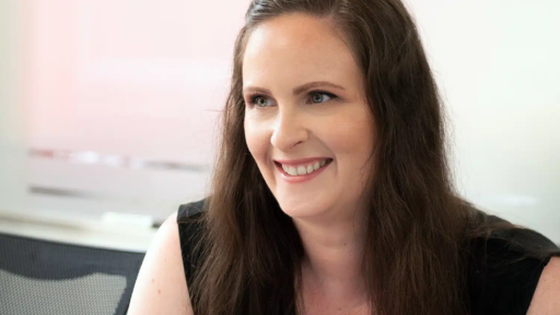 Helen Imbusi with long brown hair wearing a sleeveless black top, seated indoors at a desk or table. The background shows a light-colored wall and part of an office chair, suggesting a professional setting.