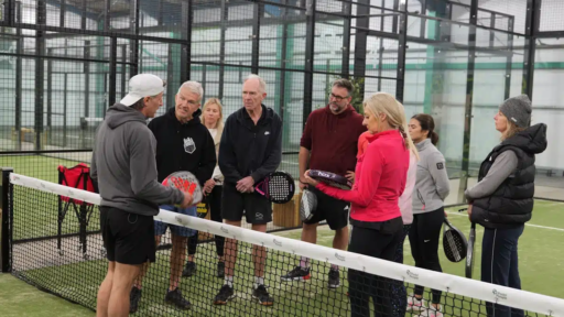 A group of people standing on an indoor padel court near the net, holding padel rackets. They appear to be listening to an instructor who is demonstrating with a racket. The court is enclosed by glass walls and metal fencing, with bright overhead lighting and visible “Padel People” branding.