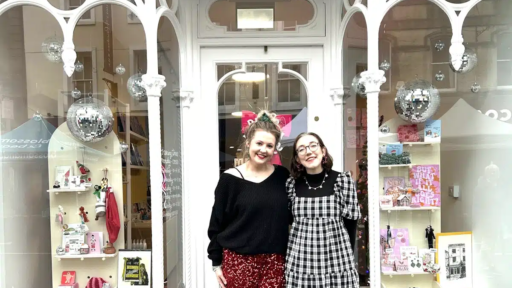 Two people standing in front of the Pixie and Pickle shop entrance, framed by ornate white window arches. The shop display features colourful gifts, books and decorative items, with silver disco balls hanging inside the windows.