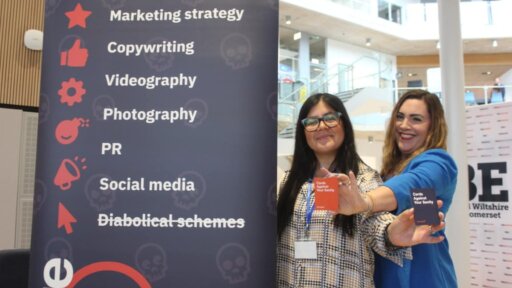 Jess Edwards (left) and Sara Witham (right) from Evil Empire marketing stand beside a branded banner at TBE Show 2025. The banner lists services including Marketing strategy, Copywriting, Videography, Photography, PR, Social media, and Diabolical schemes. Both are holding business cards toward the camera, with a modern, open-plan venue visible in the background.