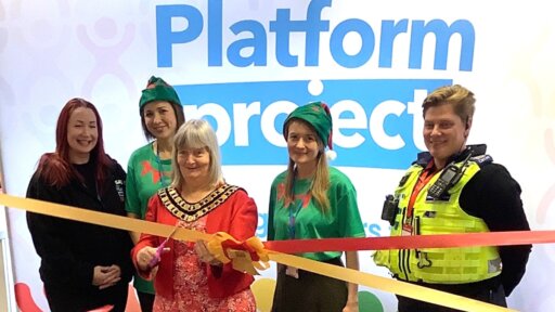 A ribbon-cutting ceremony in front of a large display that reads “The Platform Project.” Five individuals are standing together, including two wearing green elf costumes and one in a high-visibility police vest. The person in the center is holding scissors and cutting a red ribbon tied with a gold bow. The background features colorful abstract figures in shades of red, green, and yellow.