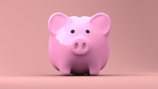 Pink ceramic piggy bank with a glossy finish, positioned on a smooth surface against a solid light pink background. The piggy bank has a rounded shape with two small ears, a snout featuring two nostrils, and black circular eyes, symbolizing savings and financial concepts.