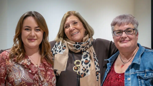 Three people standing side by side indoors, with arms around each other. From left to right: Lauren Roberts wearing a patterned maroon blouse, Janet Shreeve in a dark top with a beige leopard-print scarf and a large circular pendant necklace, and Fiona Scott in a denim jacket over a red sequined top. Neutral background.