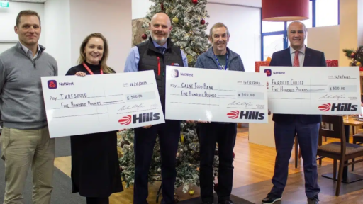 ive people stand indoors in front of a decorated Christmas tree, each holding a large ceremonial NatWest check for £500.00. The checks, issued by Hills on 16/12/2023, are made out to Threshold, Calne Food Bank, and Fairfield College, highlighting a festive charitable donation event.