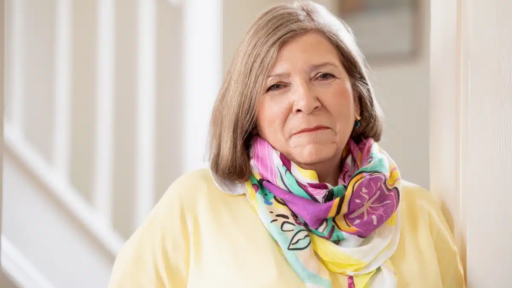 A portrait of Janet Shreeve standing indoors near a staircase, wearing a light yellow top and a colourful patterned scarf.