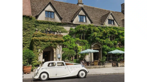 Historic stone building of The Old Bell Hotel in Malmesbury, covered in lush green ivy, with pitched roofs and dormer windows. A classic white vintage car is parked in front, and two outdoor tables with green parasols are set up near the entrance, creating a charming and romantic Cotswolds setting under a bright blue sky.