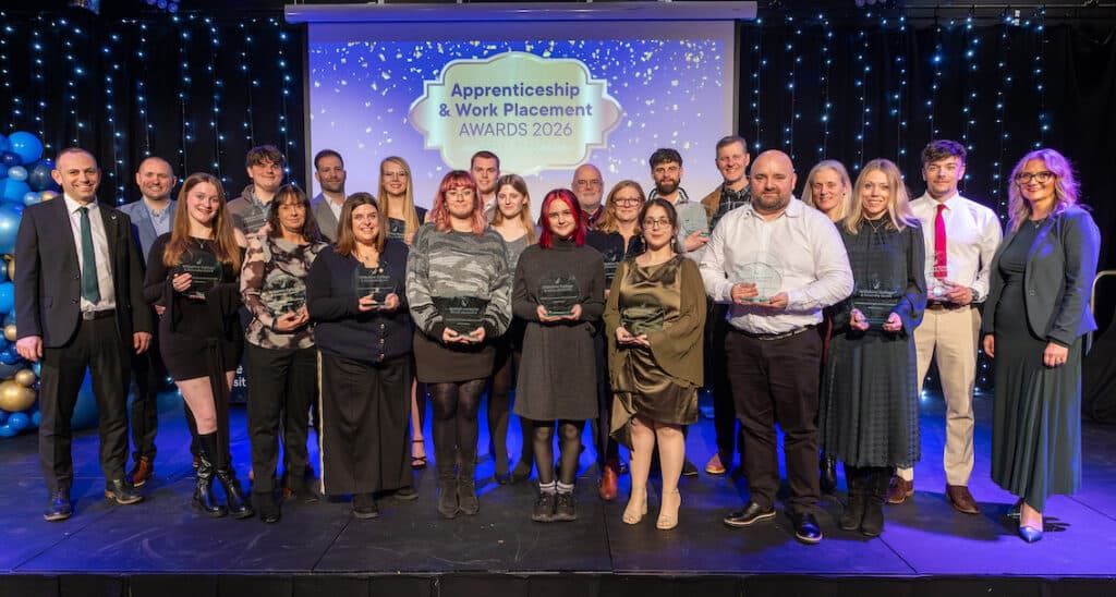 Apprentices from Wiltshire College & University Centre standing on stage holding glass awards during the annual Apprenticeship and Work Placement Awards ceremony, with event branding, balloons and lights in the background