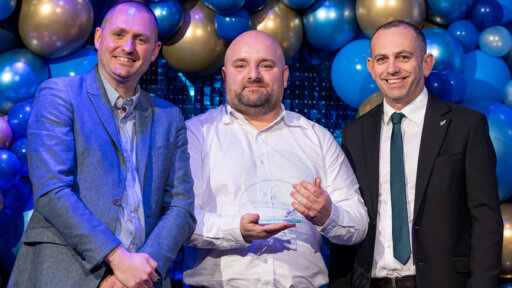 Bogdan Soporan at the Wiltshire College & University Centre’s annual Apprenticeship and Work Placement Awards with his award. He is wearing a white shirt and behind him are blue and gold balloons.