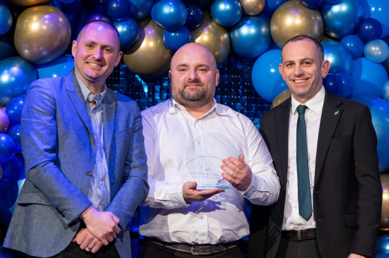 Bogdan Soporan at the Wiltshire College & University Centre’s annual Apprenticeship and Work Placement Awards with his award. He is wearing a white shirt and behind him are blue and gold balloons.