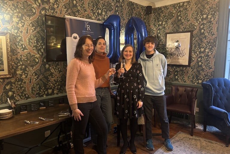 A group photo of KR HR & Training Consultancy Ltd celebrating their 10th anniversary. There is flowery green and brown wallpaper behind them