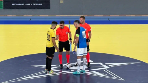 A group of four futsal players and referees stand in a circle at centre court on an indoor pitch, gathered around a ball for the pre‑match coin toss. Two players wear yellow and black kits, while the others wear blue and white. The referees are dressed in bright orange shirts. The court features a large star emblem at the centre.