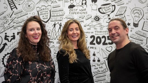 TBE's Anita, Anna and Ben stand in front of a white illustrated backdrop filled with hand‑drawn icons, awards, and motivational phrases. The person on the left wears a patterned black and pink top, the person in the centre wears a black dress with subtle shimmer, and the person on the right wears a dark long‑sleeve shirt. The backdrop features sketches such as “Best Start Up 2023,” “Best User Experience,” “Charity Not for Profit of the Year,” and various doodles related to innovation, collaboration, and awards.