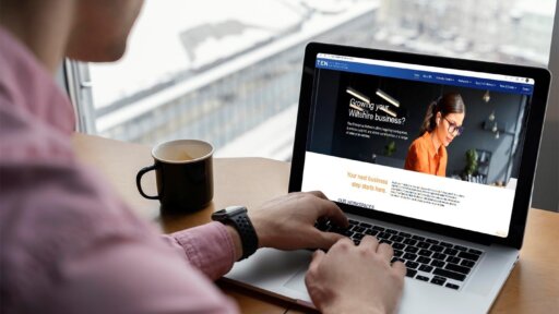 A person sits at a desk by a large window, working on a laptop that displays a business‑focused website. A coffee mug is placed to the left of the laptop, and an urban, snowy cityscape is visible outside the window.