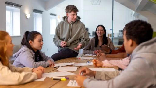 A group of people sit around a large wooden table in a bright, modern room, engaged in a collaborative discussion. Papers, pens, and highlighters are spread across the table, and one person is standing and speaking while others listen and take notes. The atmosphere appears to be focused and conversational.