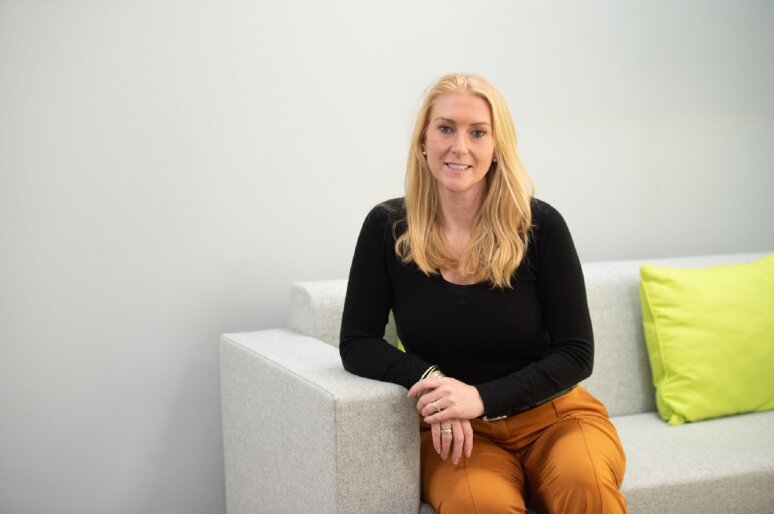 Ashlea Fisher with long light‑coloured hair is seated on a light grey sofa in a bright, minimalist room. She is wearing a black long‑sleeved top and orange trousers, with one arm resting on the sofa arm and the other on their knee. Two lime‑green cushions are placed on the sofa beside them.