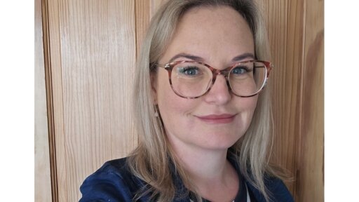 Becky Richens standing indoors in front of a wooden panelled wall, wearing a dark top with layered fabrics and shoulder‑length hair.