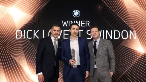 Three people in formal suits stand together on a stage at an awards event. The person in the centre is holding a trophy. Behind them is a large illuminated backdrop featuring the BMW logo and the words “WINNER DICK LOVETT SWINDON,” with geometric light patterns radiating outward.