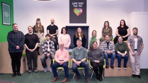 “A group photo of the GEL Studios team taken indoors in a modern office space. Fifteen people stand on stepped seating at the back, while five people sit in bright green chairs at the front. The room features green accents, circular ceiling lights, and a framed sign on the wall that reads ‘Love What You Do’ with a colourful heart graphic. Everyone is posed facing the camera.