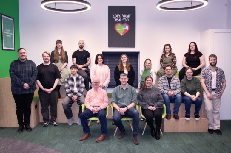 “A group photo of the GEL Studios team taken indoors in a modern office space. Fifteen people stand on stepped seating at the back, while five people sit in bright green chairs at the front. The room features green accents, circular ceiling lights, and a framed sign on the wall that reads ‘Love What You Do’ with a colourful heart graphic. Everyone is posed facing the camera.