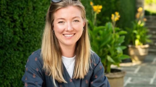 Georgina Riches with long, light-coloured hair is seated outdoors in a garden setting. They are wearing a dark patterned shirt over a light top, with sunglasses resting on their head. Behind them is a neatly trimmed hedge, potted plants, and stone steps leading upward, all in bright natural light.