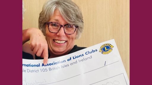 Jane Boulton holding a large ceremonial cheque from the International Association of Lions Clubs, showing the British Isles and Ireland district branding. A hand points toward the cheque, which displays an amount written as £5,000. The background consists of a light wooden surface.