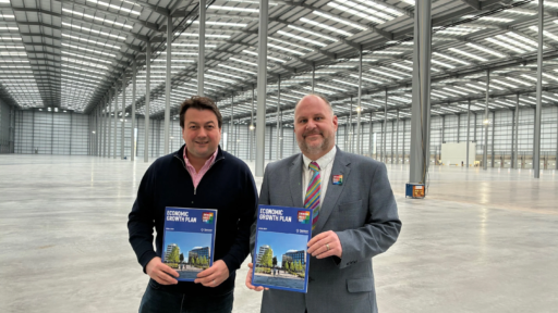 Cllr Robbins with James Watson standing inside a large empty warehouse at Panattoni Park holding copies of the Swindon Economic Growth Plan booklets, with high ceilings, skylights, and concrete flooring visible in the background.