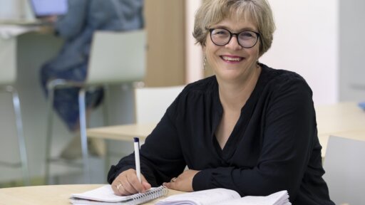 Jo Smyth is seated at a desk in a bright indoor workspace, writing in an open notebook with a pen. Another open notebook lies on the desk in front of her. In the background, another person sits on a high chair working at a laptop near a counter. The setting has light-coloured furniture and a soft, modern atmosphere.