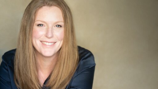 Kellie Chandler ith long straight hair is seated indoors against a softly lit neutral background. She is facing the camera with a relaxed posture, wearing a dark, smooth-textured top