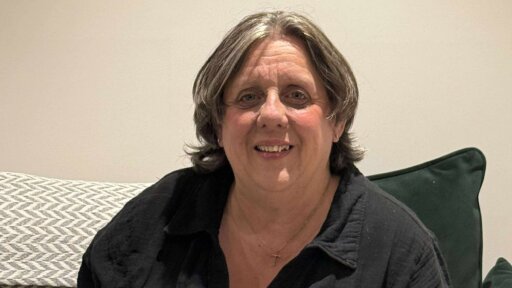 Lisa Jackson wearing a dark button‑up shirt and a small cross necklace is seated indoors on a sofa with patterned and dark green cushions against a light‑coloured wall.