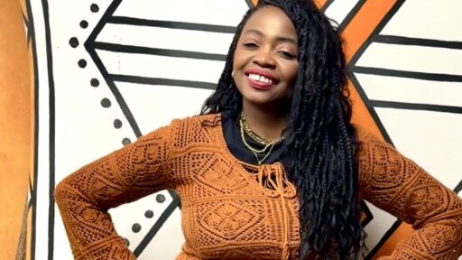 Lydia Moore standing in front of a bold geometric mural with black, white and orange patterns. The person is wearing an orange textured top with long sleeves and has long braided hair over one shoulder. She is smiling and standing with hands on hips.