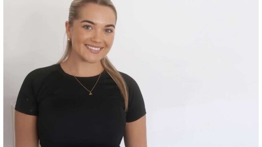 Paige Cook wearing a black short‑sleeved top and a delicate gold necklace, seated against a plain white background.