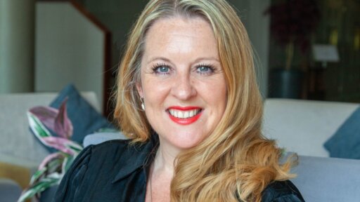 Portrait of Rachael Matwiejczyk-Booth seated indoors on a light-coloured sofa, with long wavy blonde hair and a black top, with soft furnishings and plants visible in the background.