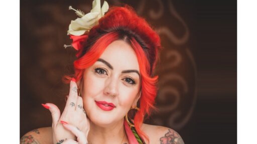 Raye Leonard with bright red, styled hair is shown against a dark patterned background. A large cream‑coloured flower accessory is positioned in the hair. The person’s hand is raised near their face, showing long red nails and several tattoos. The person is wearing a halter‑style top with visible tattoo artwork across the shoulders and upper chest.