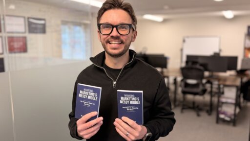 Rob Curtis stands indoors in an office setting, holding two identical books titled “Managing Marketing’s Messy Middle”. He is wearing a dark quarter‑zip top and a ring on one hand. The background shows desks, office chairs, a whiteboard, and soft overhead lighting.