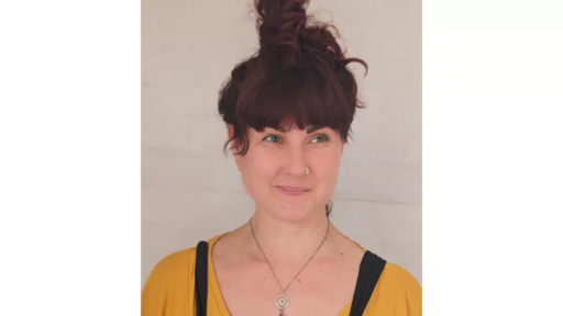 Samtosha Chi with dark reddish hair tied in a high, messy bun stands against a pale wall. They are wearing a mustard-yellow top with a black strap over one shoulder and a necklace featuring a circular charm above a purple pendant.