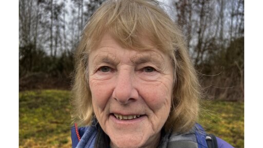 Sue Webber outdoors wearing a light jacket, standing in a grassy area with trees and winter foliage in the background.