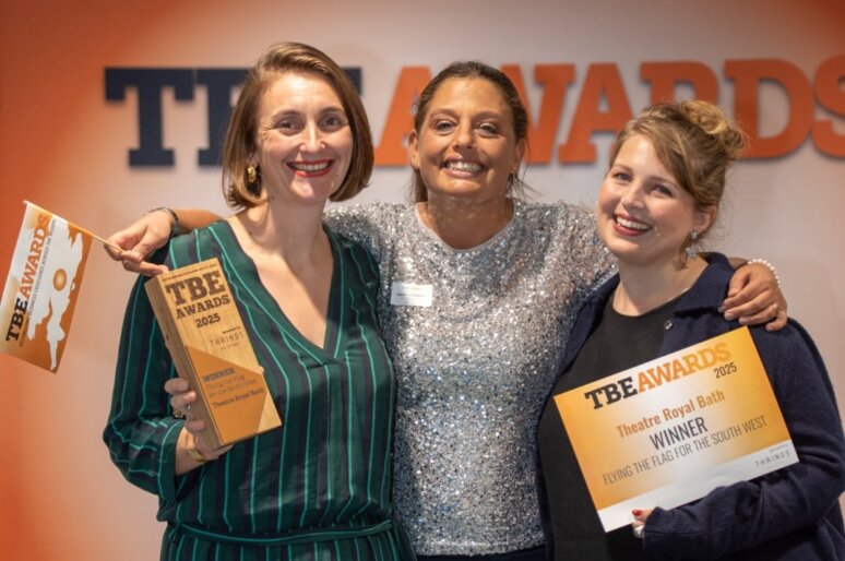 Three people standing together at an awards event, smiling and holding trophies and certificates. The backdrop reads ‘TBE Awards’. One person holds a TBE Awards 2025 trophy, and another holds a certificate announcing Theatre Royal Bath as the 2025 winner for ‘Flying the Flag for the South West.