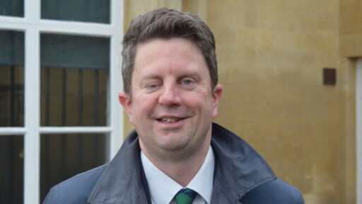 Chris Van Roon from TransWilts standing outdoors in front of a stone building with a white‑framed window behind them. The person is wearing a dark overcoat over a white shirt and a striped green-and-blue tie. The scene appears to be taken in daylight, with the architectural details of the building visible in the background.